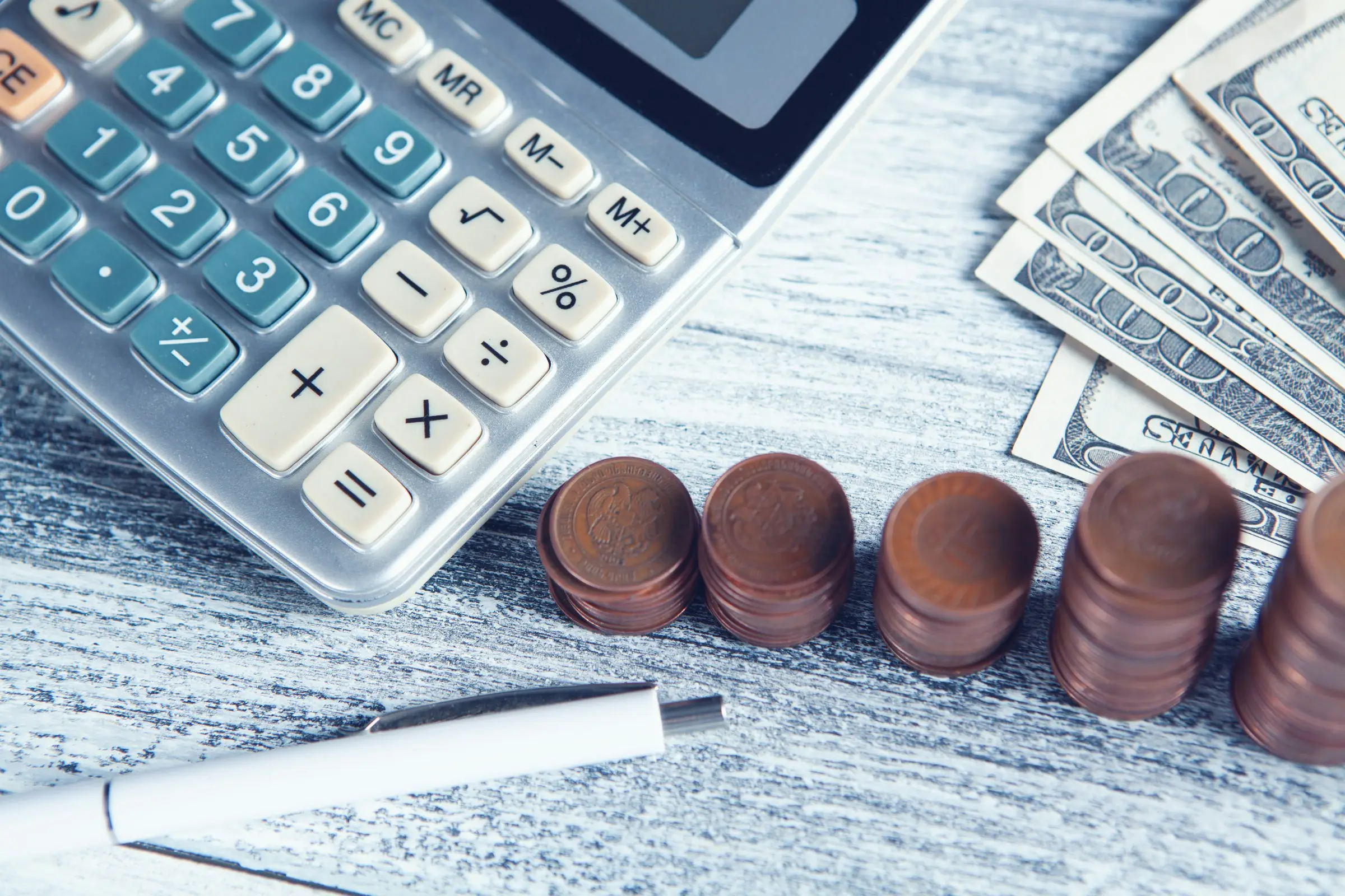 A desk calculator, a white pen, rising stacks of copper coins and a fan of banknotes on a pale wooden table.