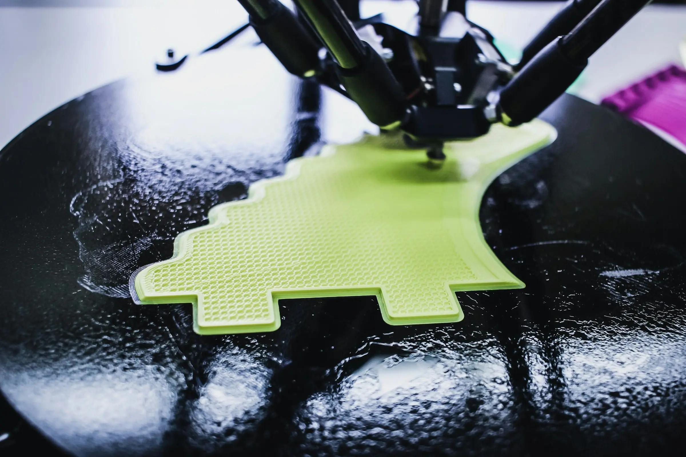 A delta-style 3D printer head on three arms printing a flat lime green shape on a glossy round black bed.