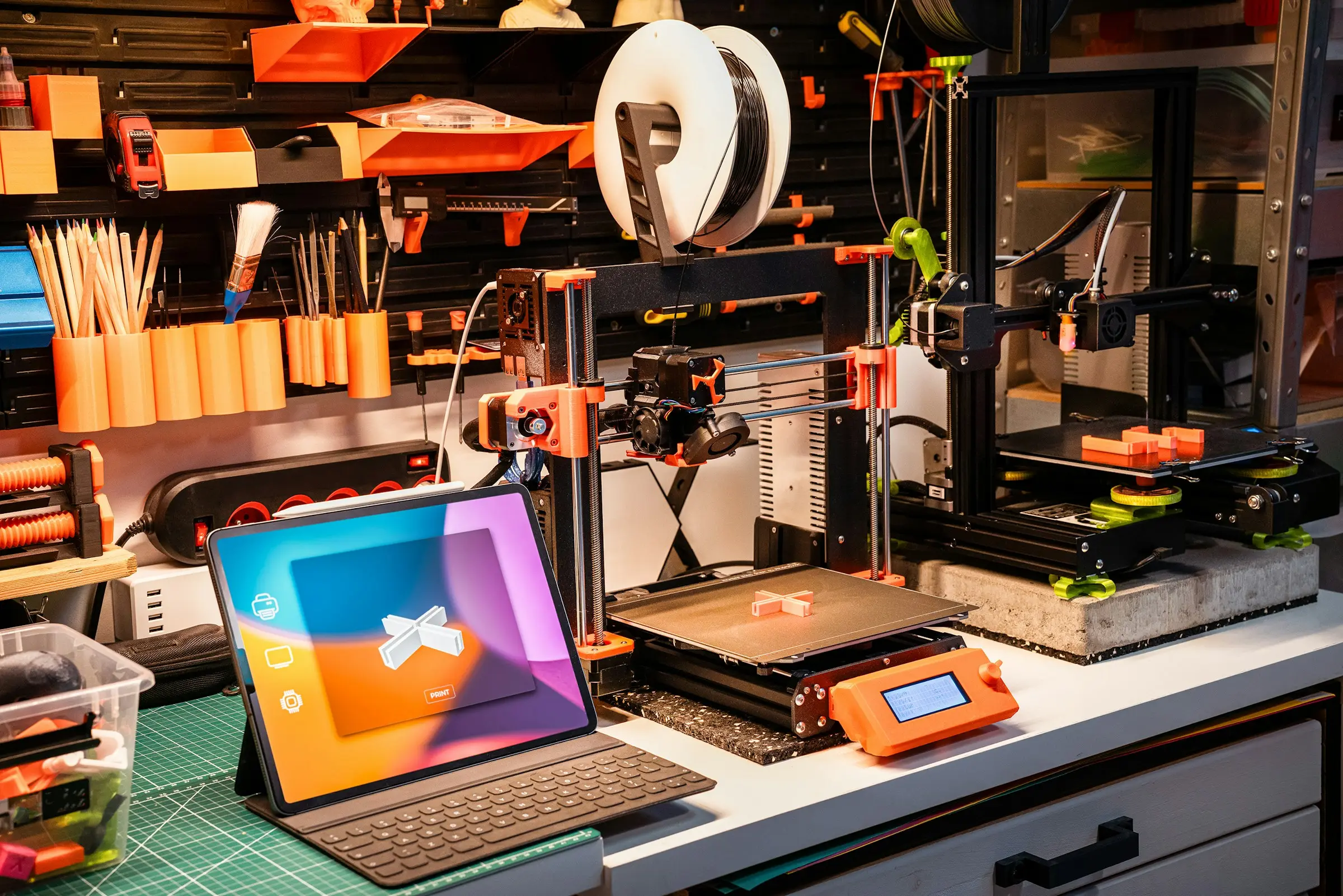 A desk with two 3D printers, a tablet with a keyboard cover and a pegboard wall of tools and orange holders.