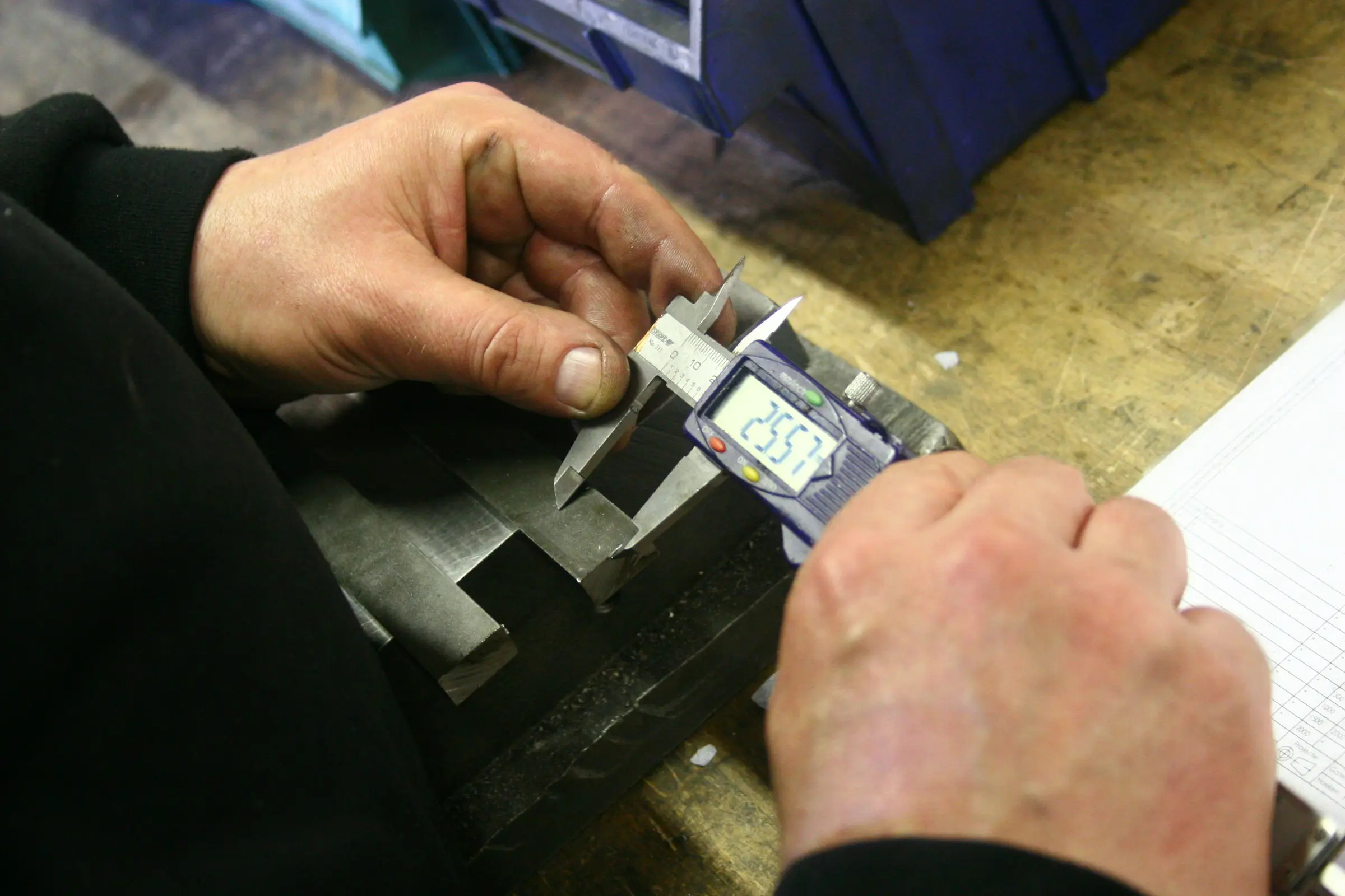 Two hands holding a digital caliper against a metal part on a workbench, the display reading 25.57.