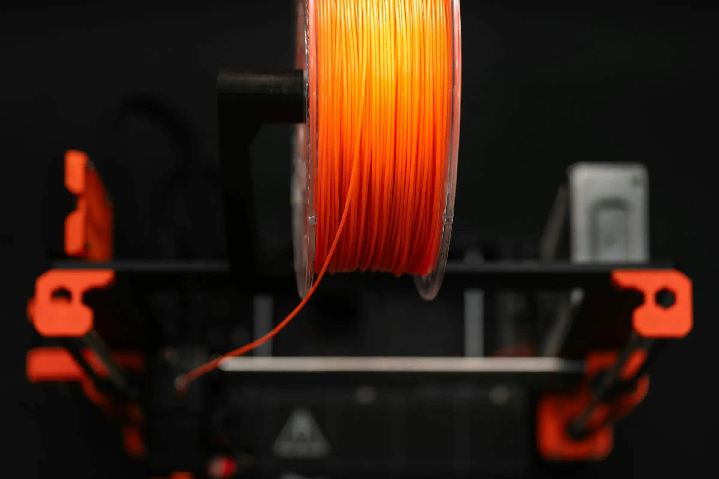 A spool of orange filament seen edge-on, mounted on top of a 3D printer against a dark background.