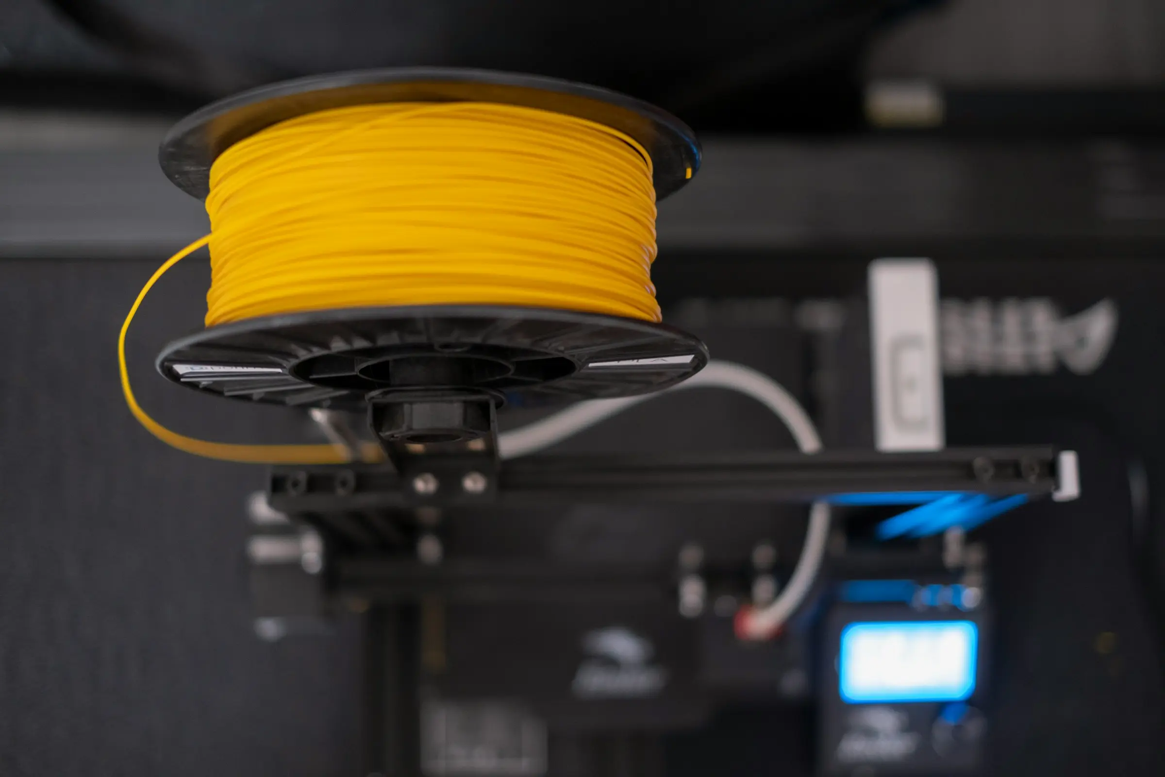A spool of yellow filament on a holder above a 3D printer, with the printer's blue screen out of focus below.