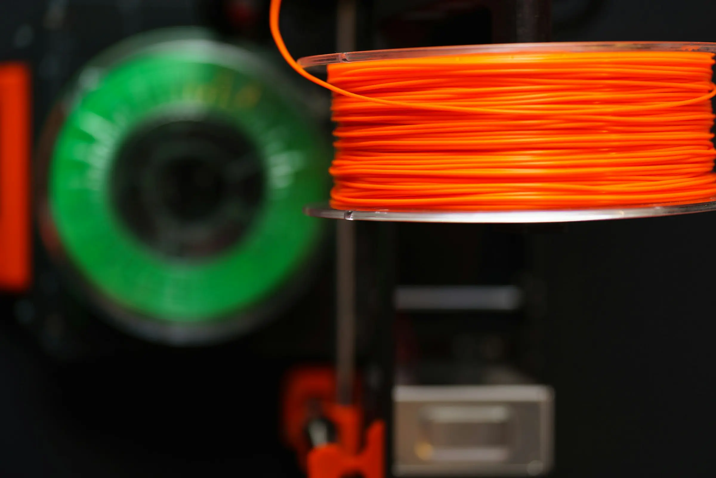Close-up of tightly wound orange filament on a clear spool, with a green spool blurred behind it.