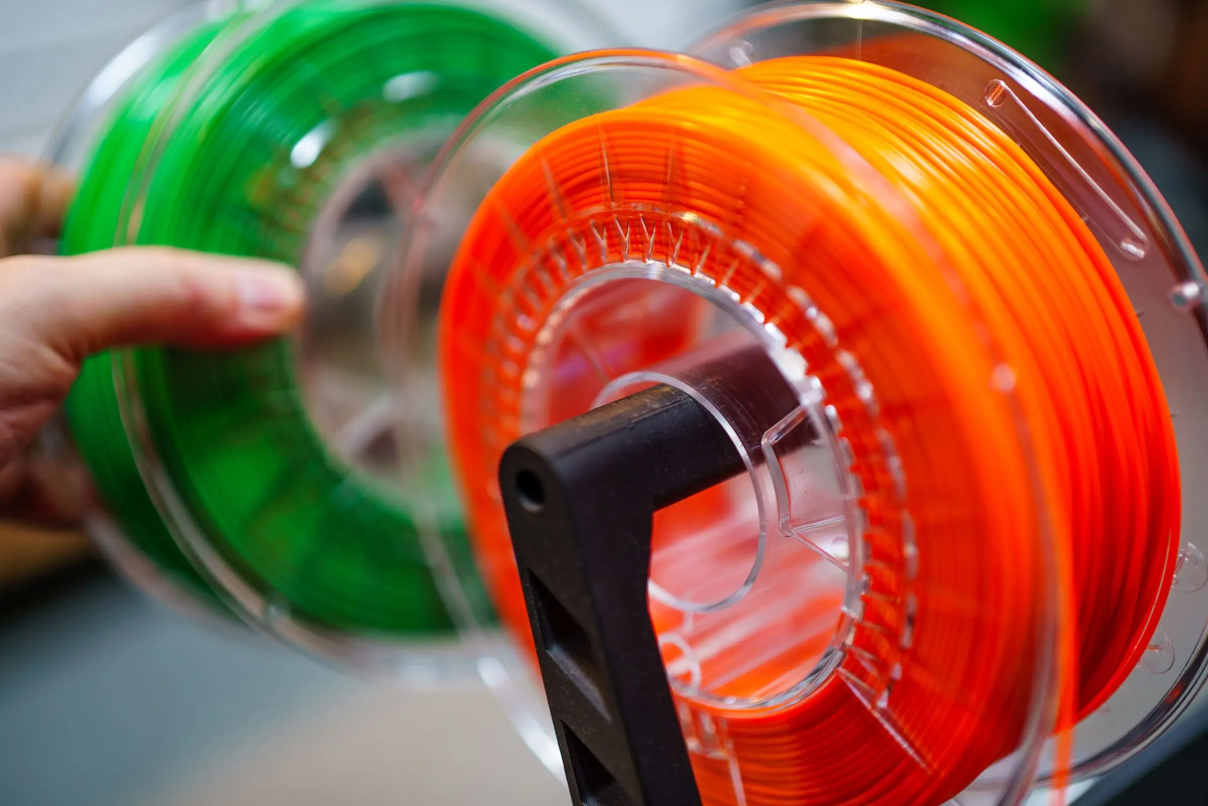 A hand steadying two clear filament spools, one orange and one green, on a black spool holder.