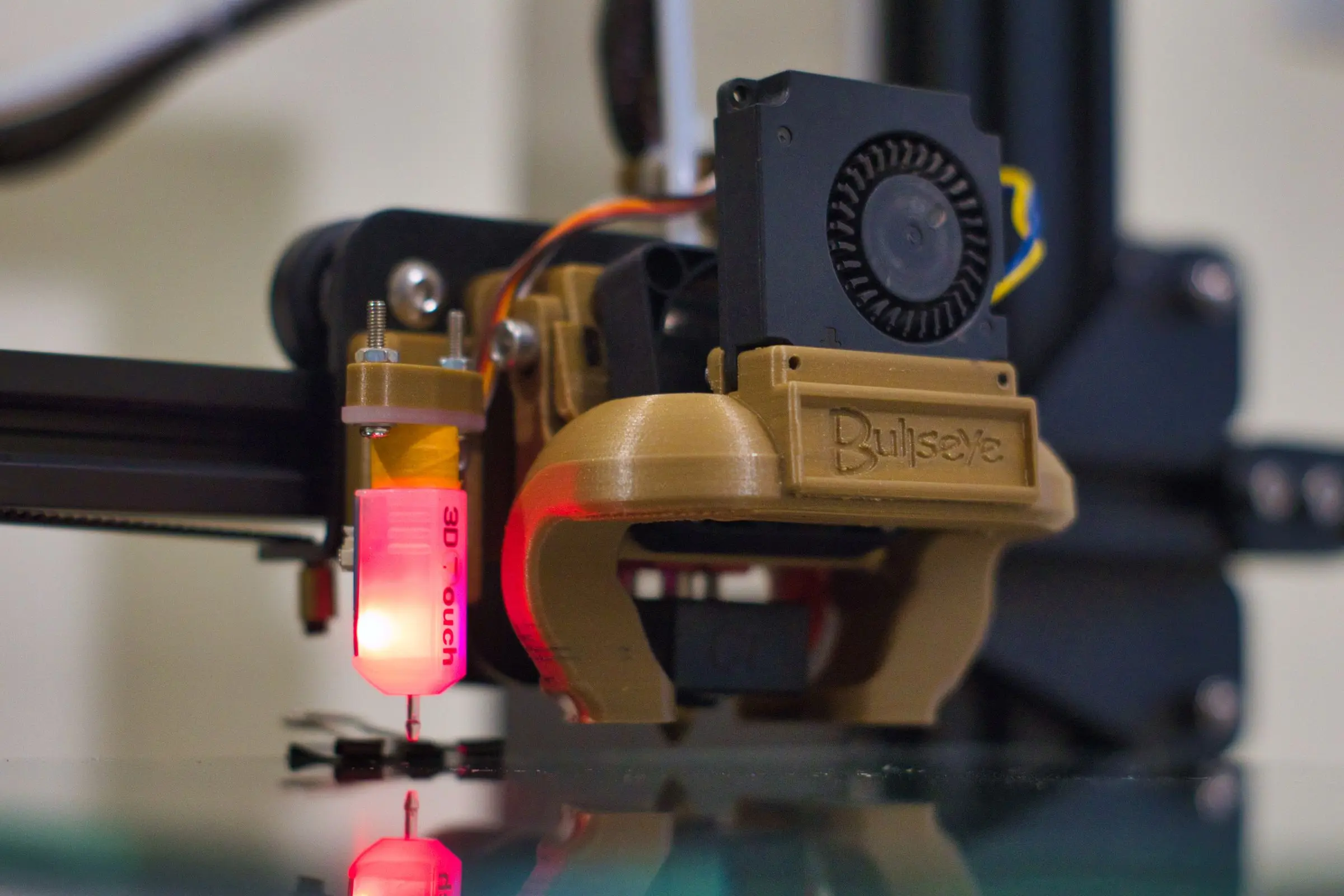 Close-up of a 3D printer print head with a printed fan duct and a bed leveling probe glowing red.