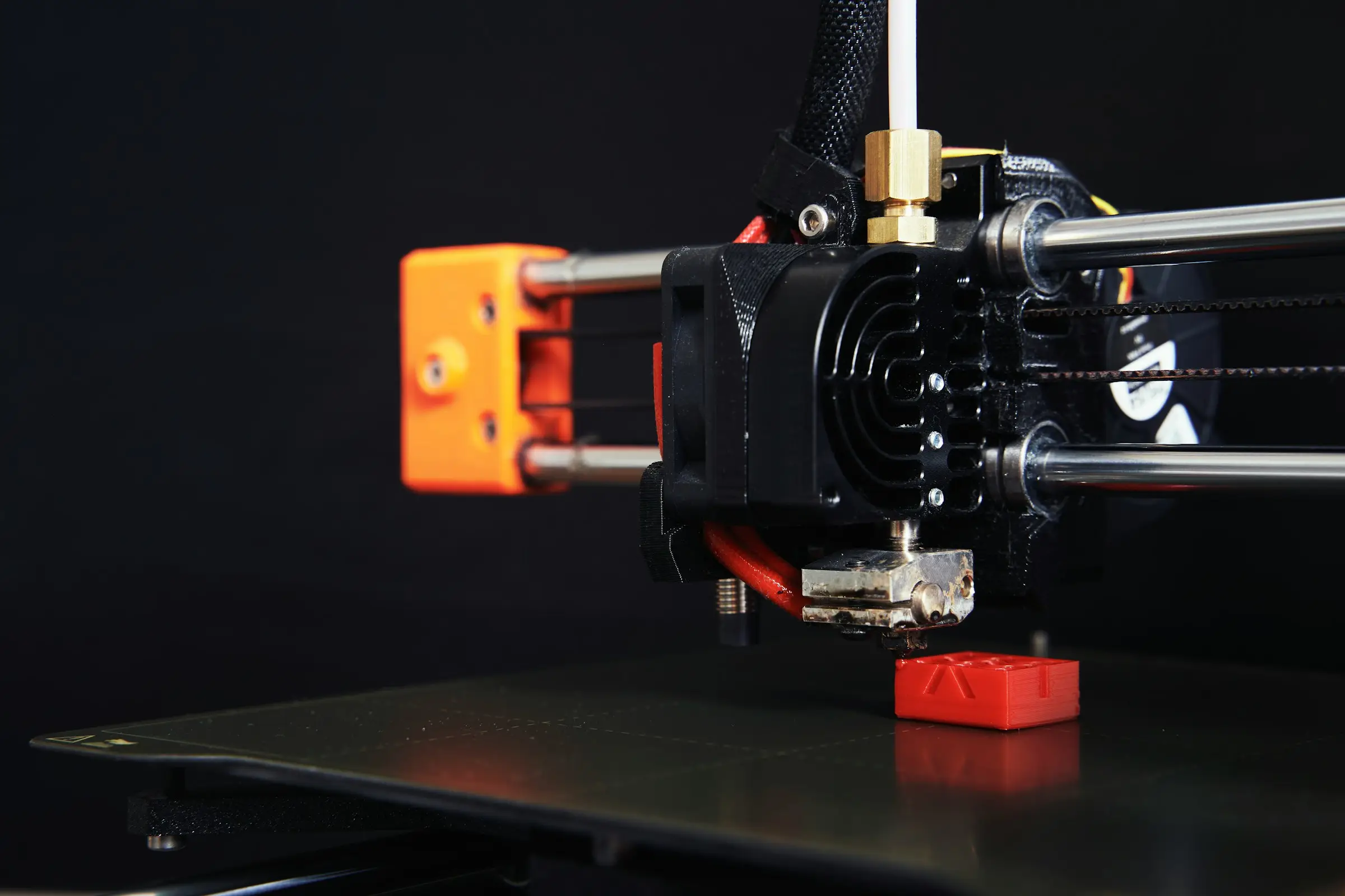 Close-up of a 3D printer hotend and heater block above a small red printed part, against a black background.