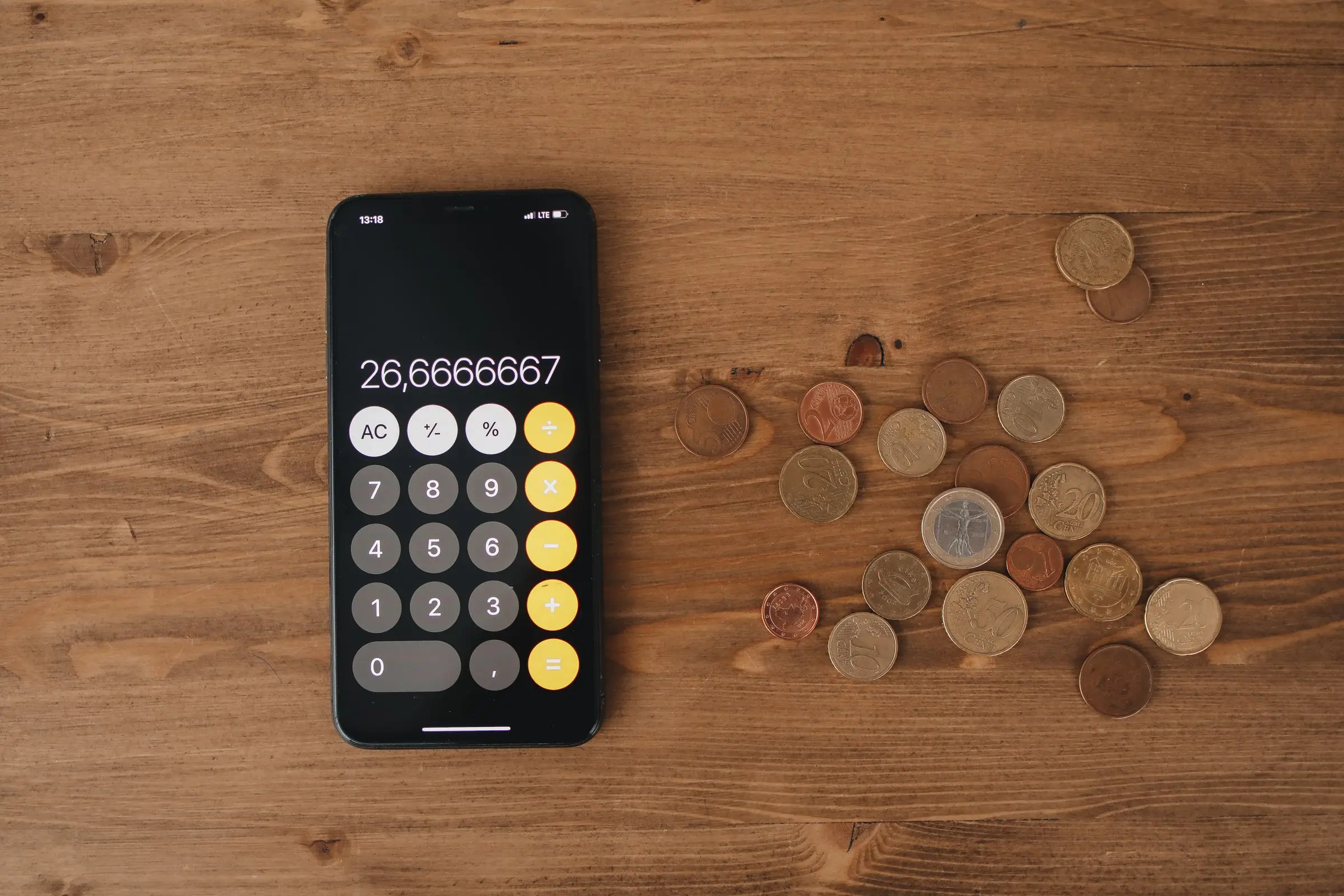 Top-down view of a phone showing a calculator app next to a scatter of loose coins on a wooden table.