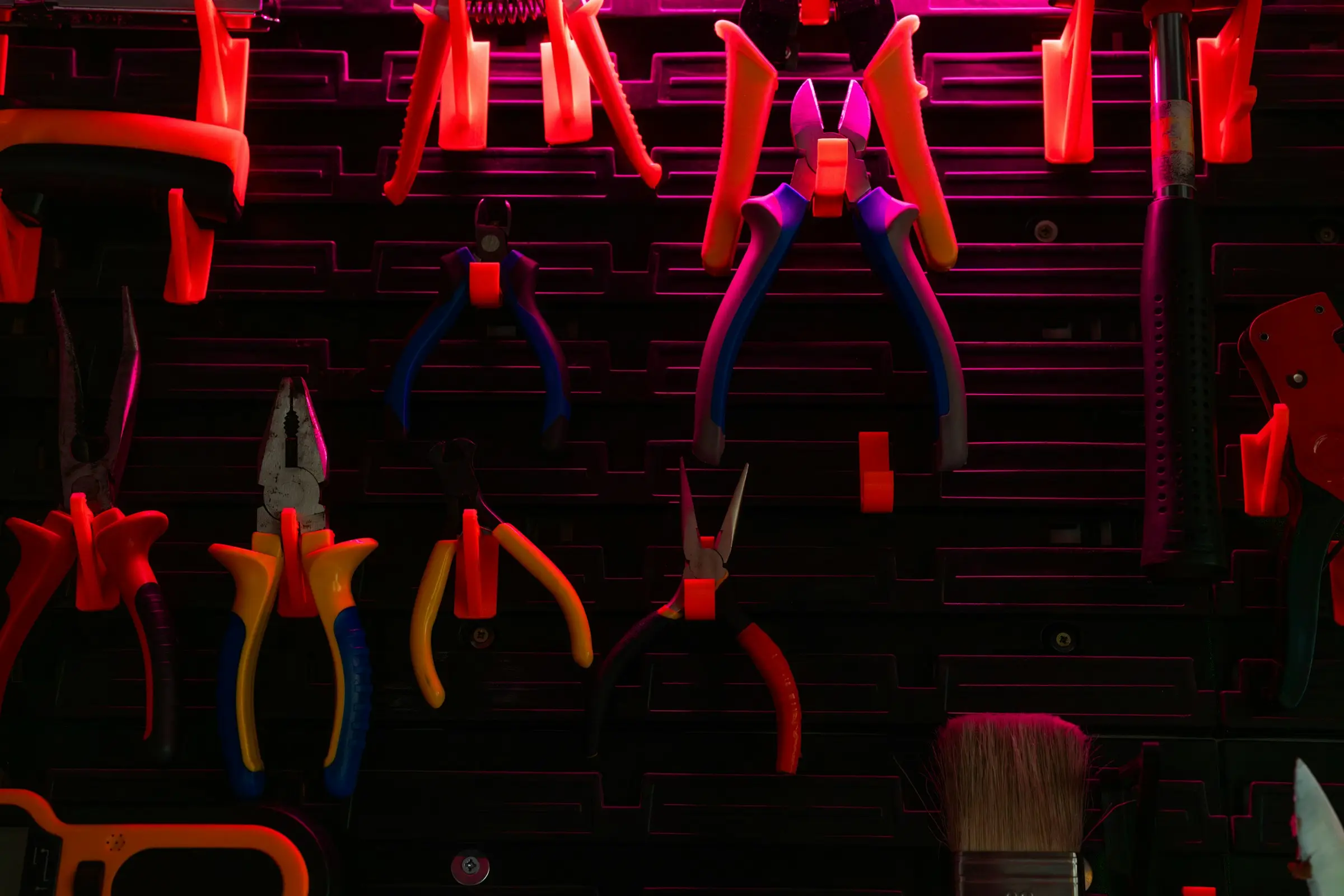Pliers, side cutters and a brush hanging on a dark tool wall with bright orange holders, lit in pink.