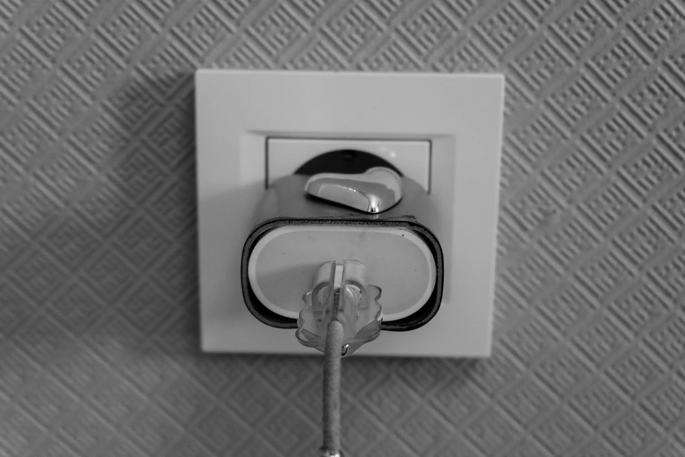 Black and white photo of a plug pushed into a wall socket on textured wallpaper, seen from the cable end.