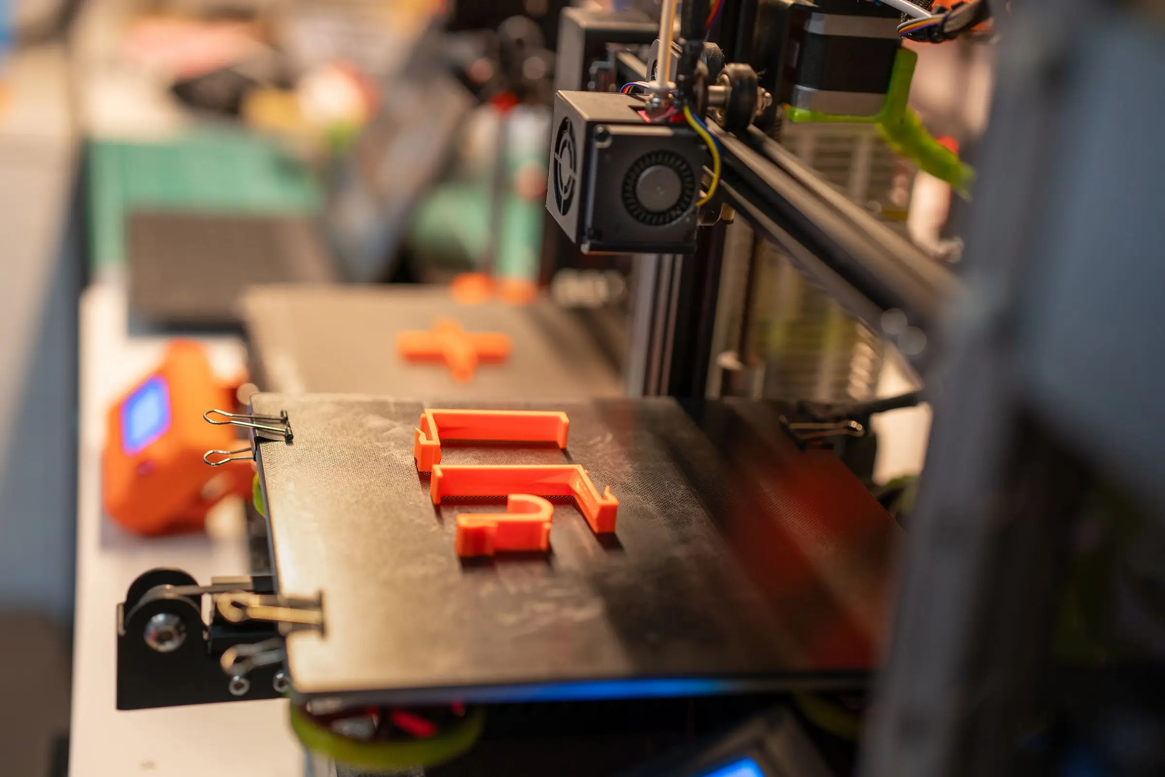 Three finished orange bracket-shaped parts sitting on a 3D printer bed held with binder clips.