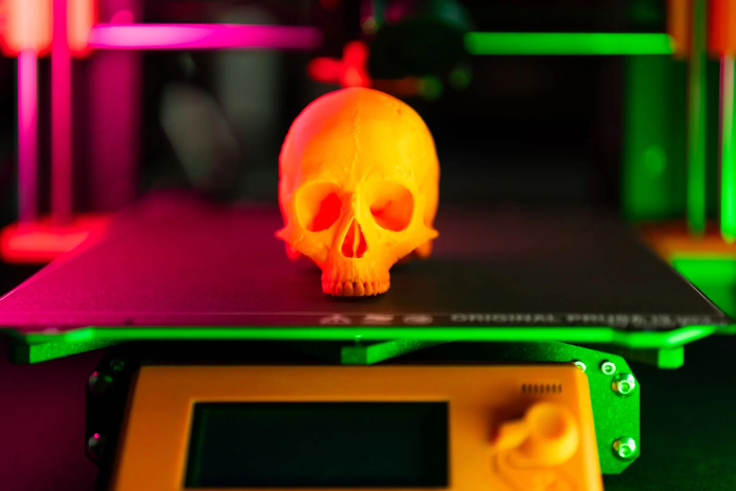 A 3D printed orange skull sitting on a printer bed, lit in pink and green.