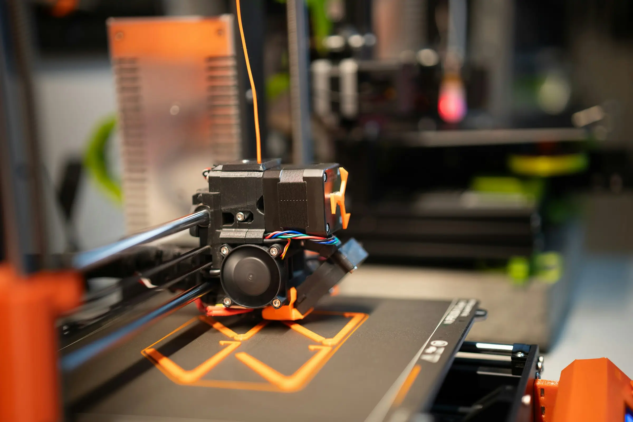 A 3D printer laying the first layer of an orange outline on a dark bed, with orange filament feeding in from above.