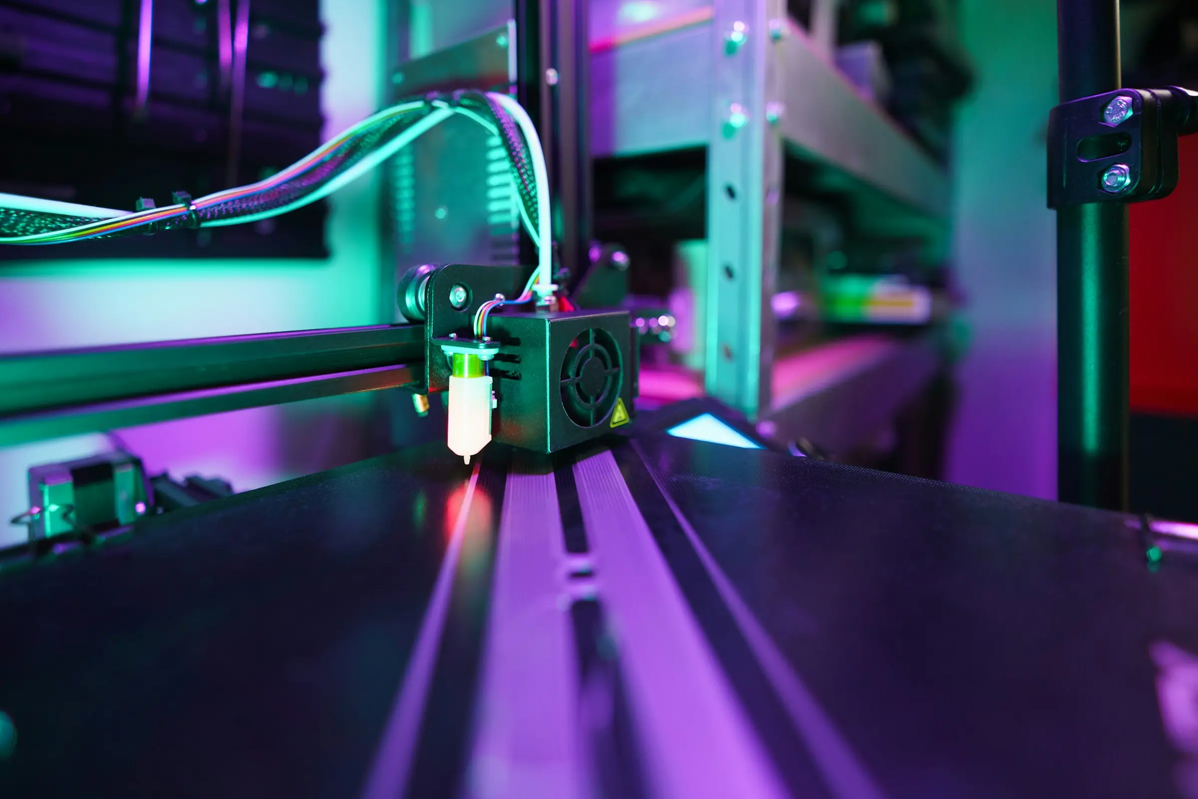 Low view along a 3D printer bed towards the print head, lit in purple and green.