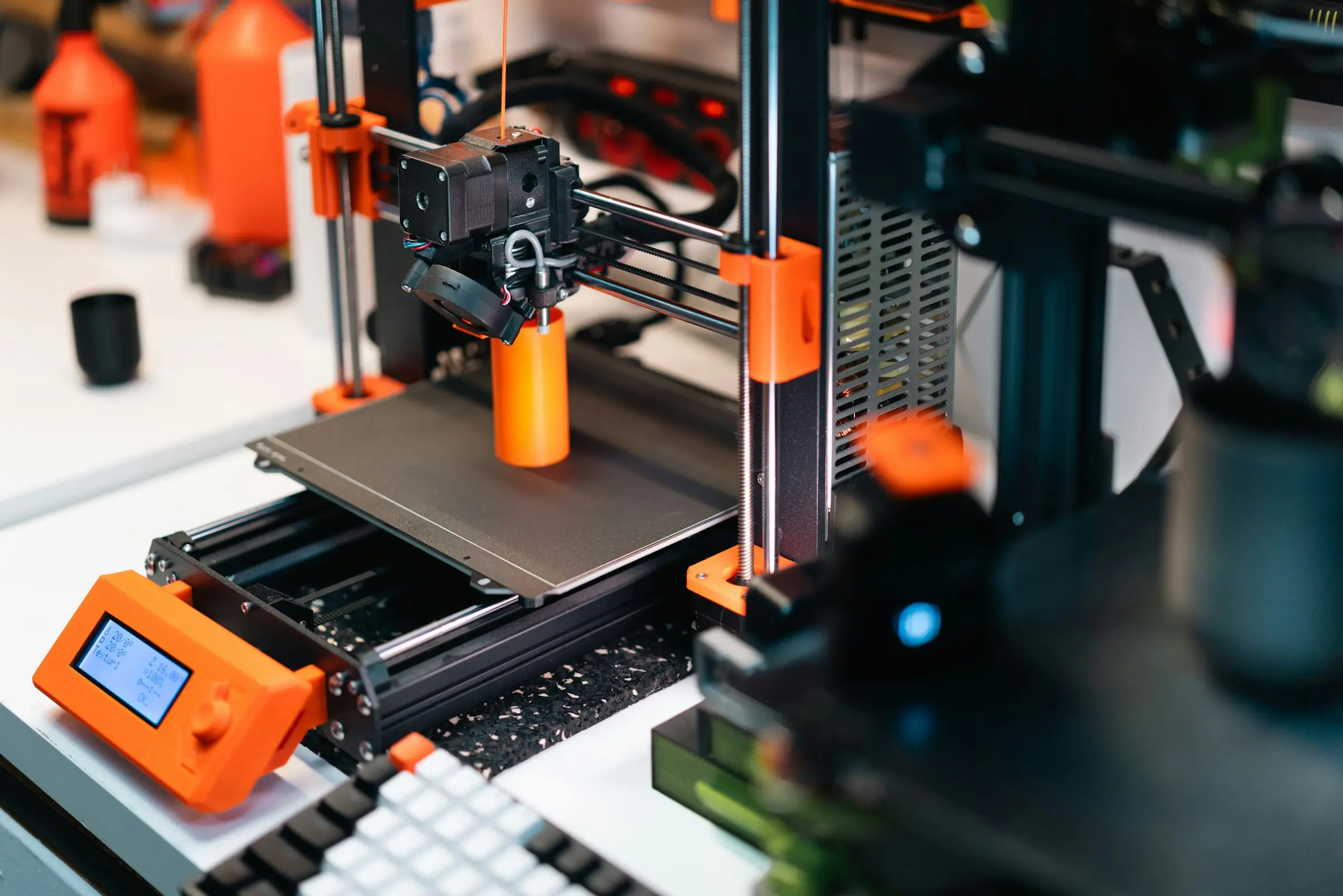 An open-frame 3D printer printing a tall orange cylinder on a desk, with a keyboard and spare bottles nearby.