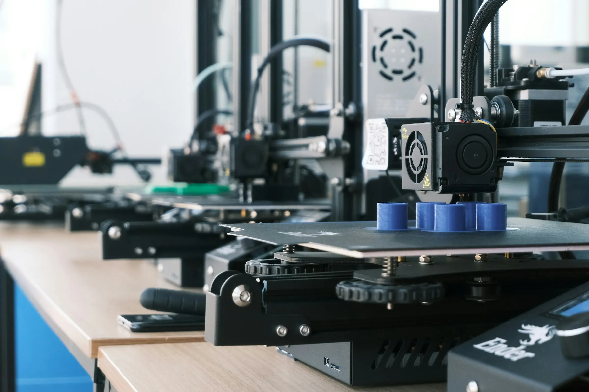 A row of open-frame 3D printers on a wooden workbench, the nearest one printing several blue cylinders.