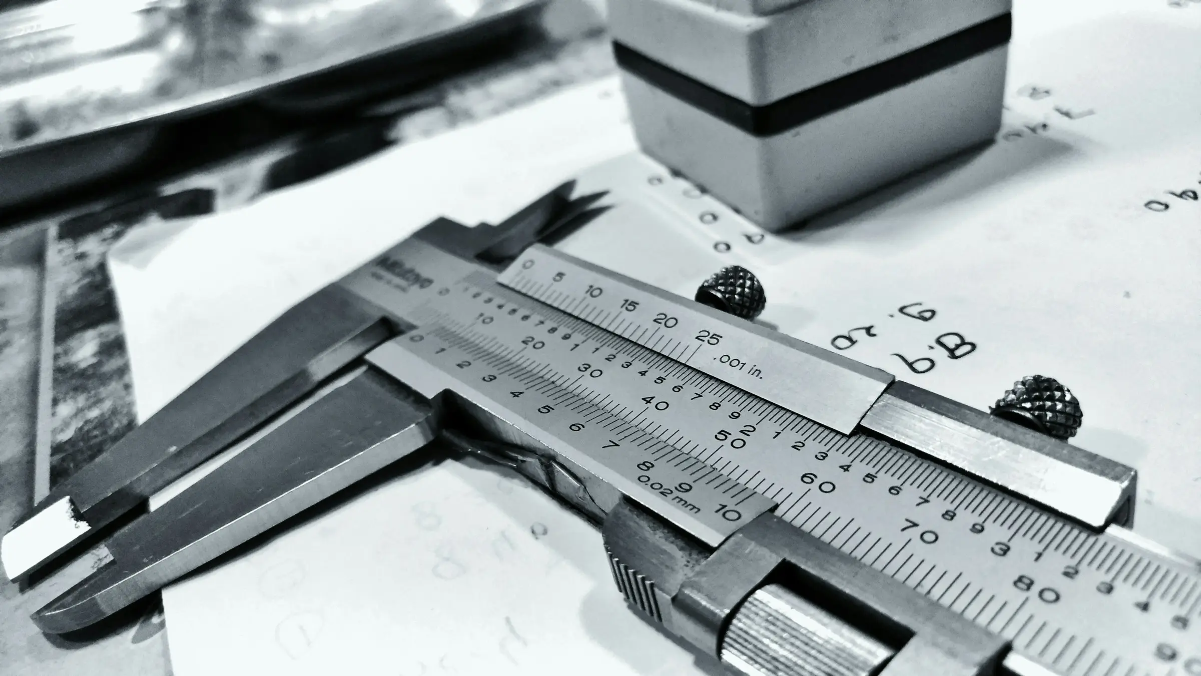 Black and white close-up of a steel vernier caliper resting on a sheet of handwritten measurements.