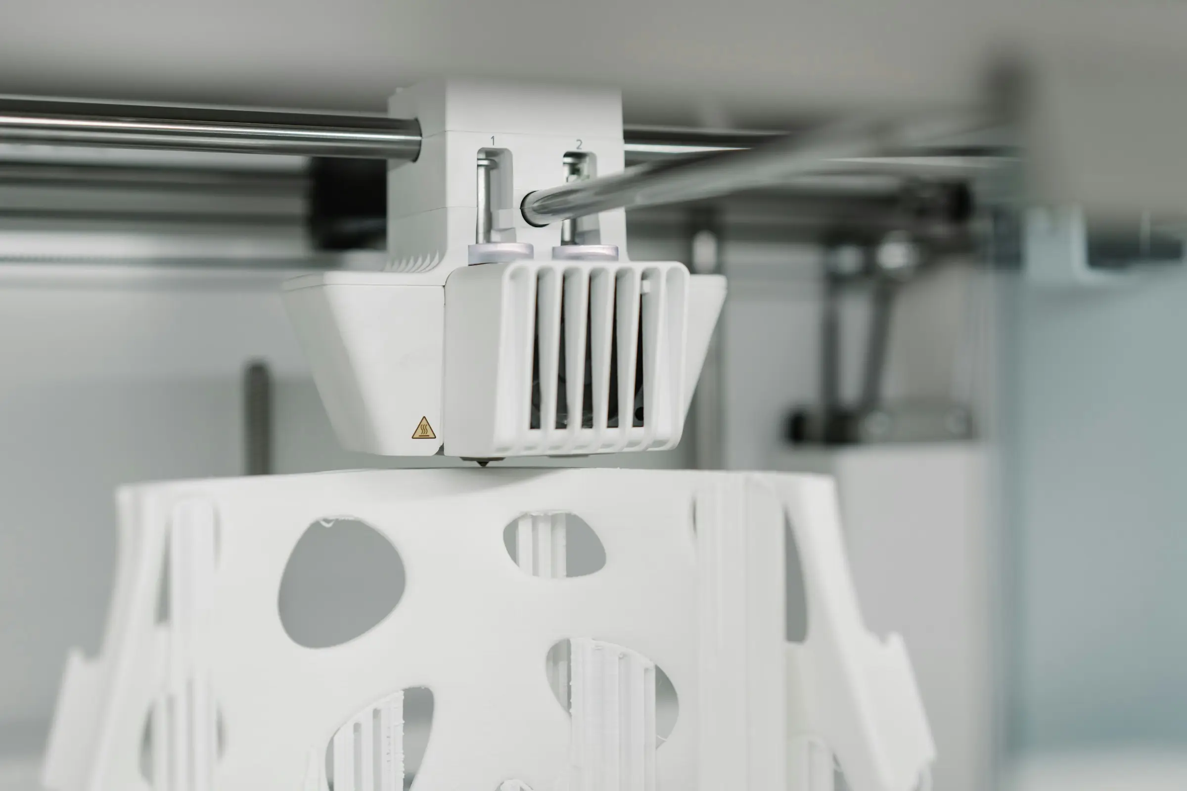 A white dual-nozzle print head working on a large white part with oval cut-outs inside an enclosed printer.