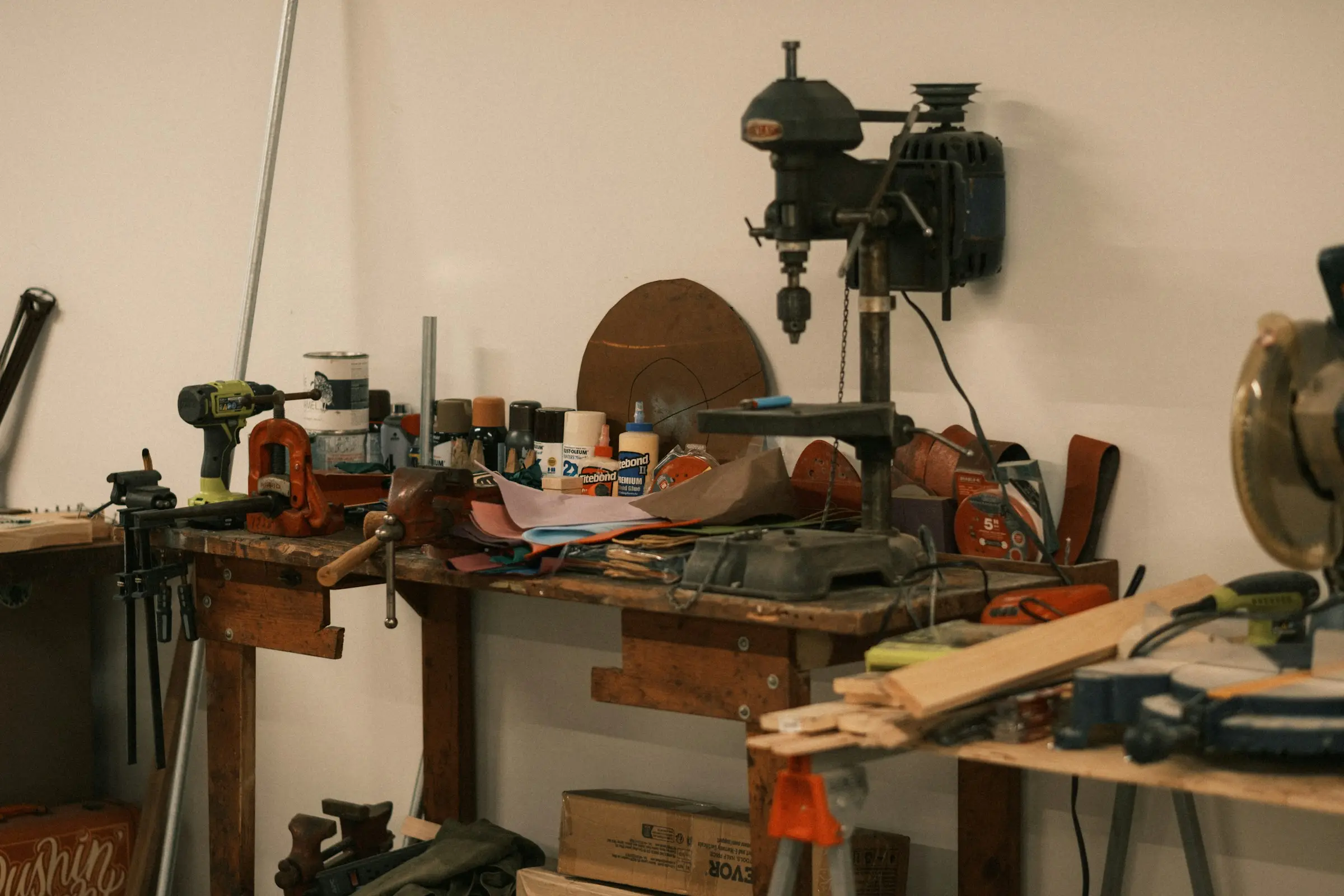 A cluttered workshop bench with a drill press, a cordless drill, clamps, glue bottles and offcuts of wood.